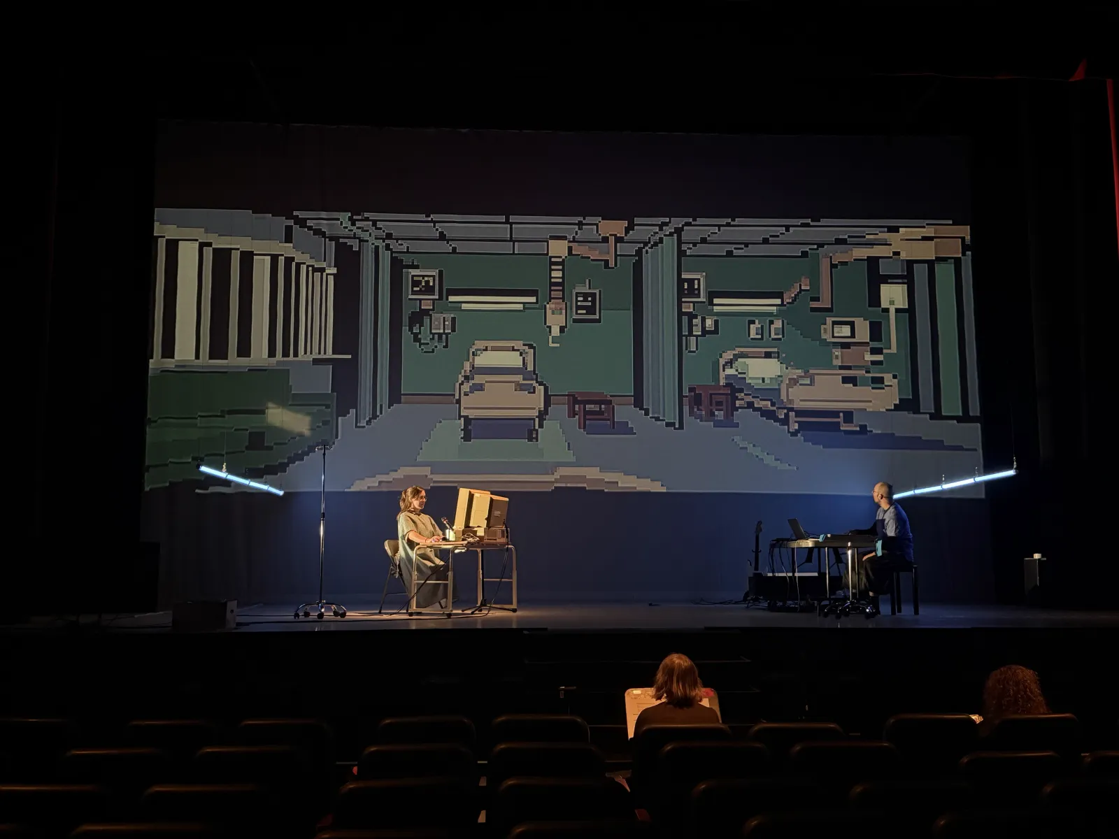 2021: a pixel-art hospital room projected over the stage, the player at an old computer on the left, the musician on the right