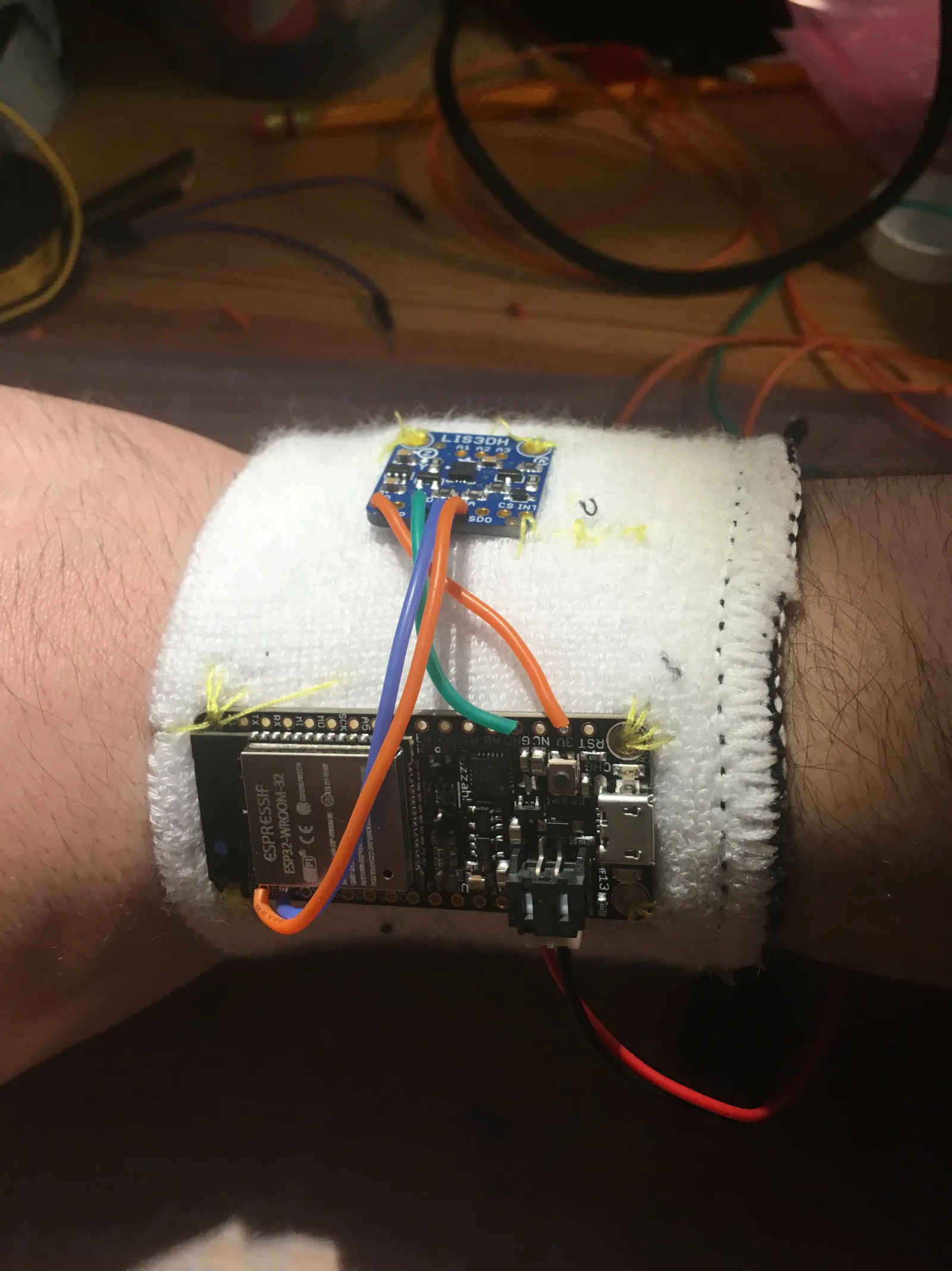 A white wristband on a wrist, with a small accelerometer board and an ESP32 board sewn on with yellow thread and joined by three wires