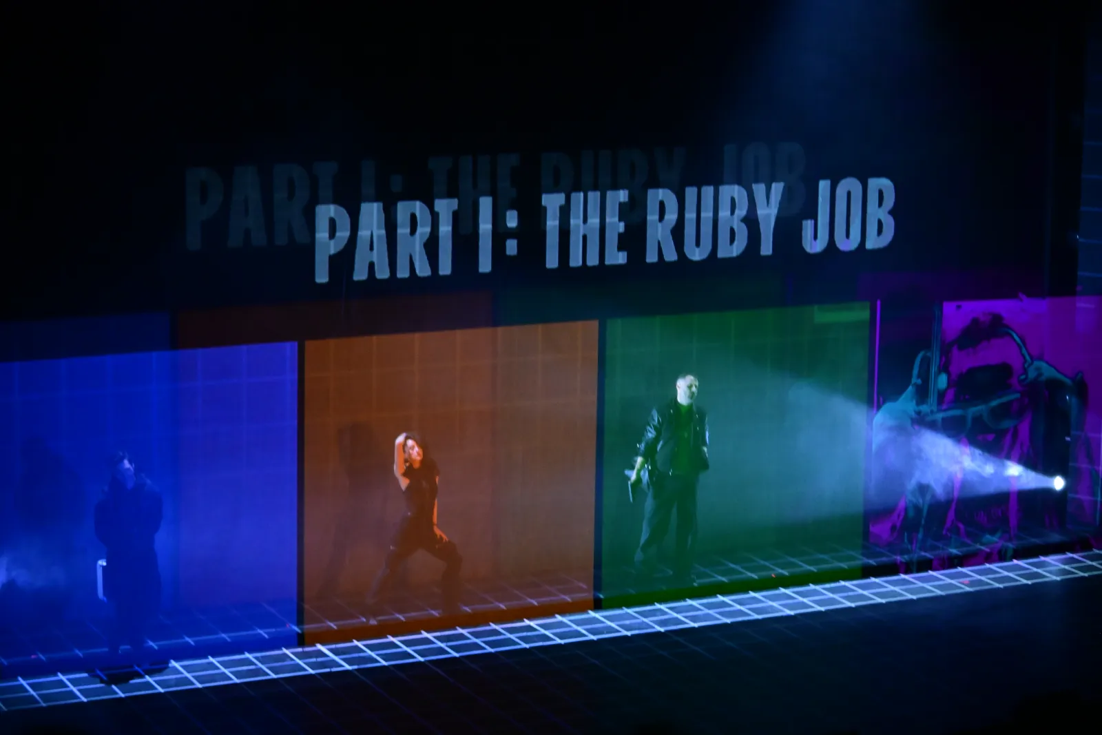 Heist: “Part I: The Ruby Job” projected above four coloured panels with performers inside them, a grid of light on the floor