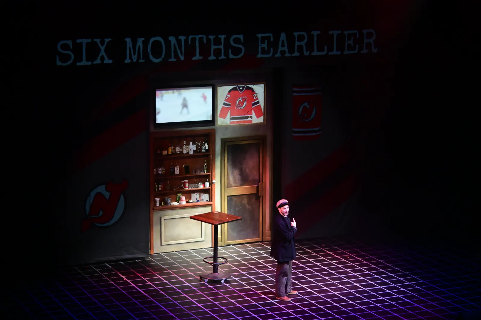 Heist: “Six months earlier” projected over a bar with a hockey jersey on the wall, an actor on the gridded floor