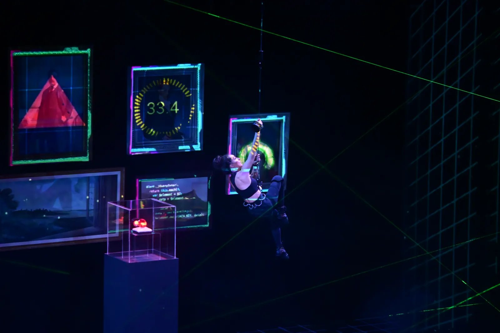 Heist: an aerialist hangs among glowing projected frames, a red triangle in one of them, lasers across the room