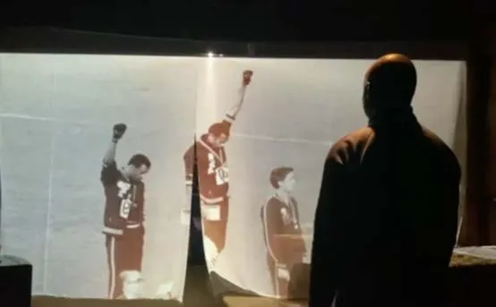 The Mountaintop: a man in silhouette watches a photograph of athletes raising their fists on a podium, projected across a hanging screen